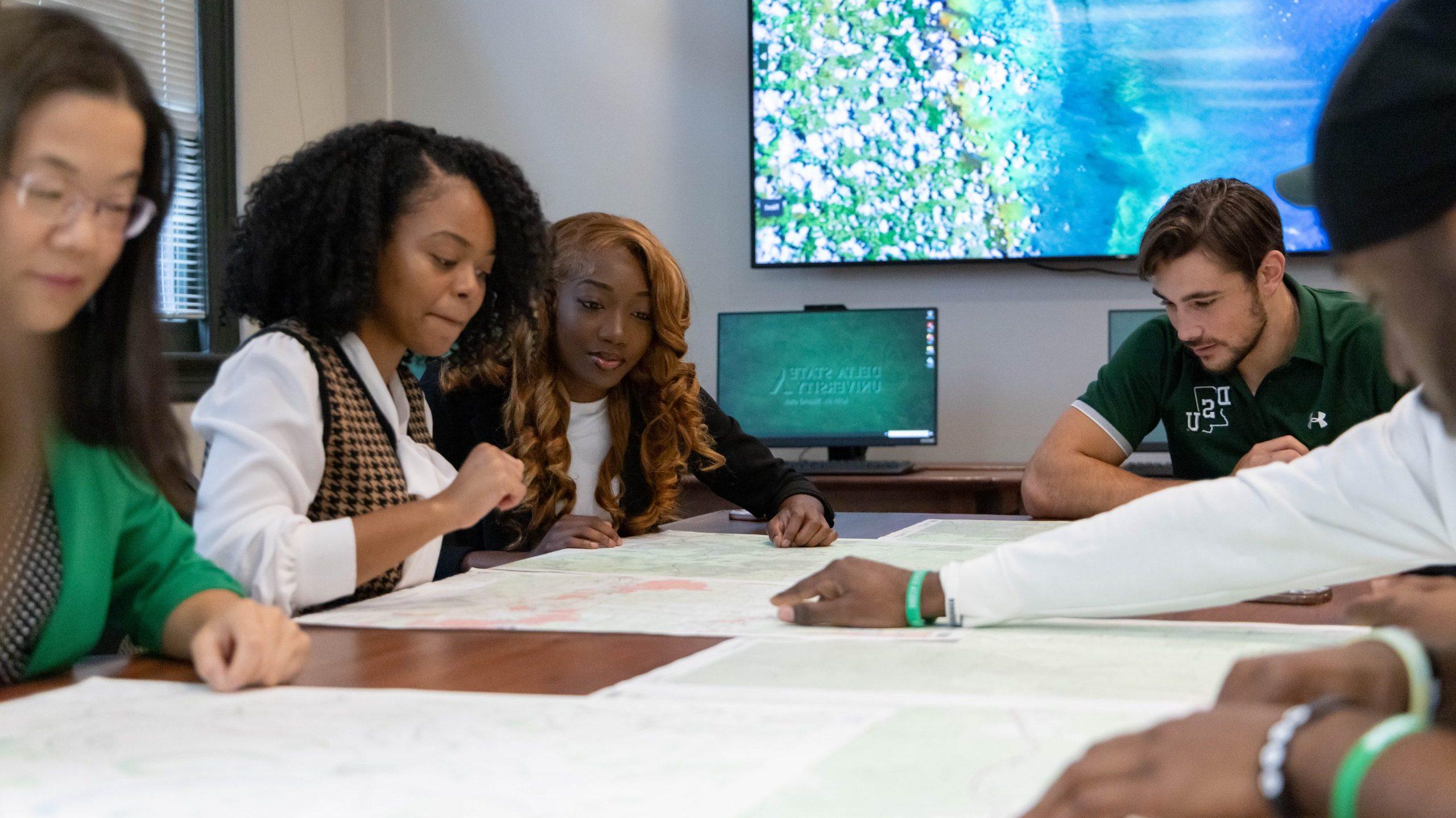 Four students and a professor sitting around a large table, looking at maps.